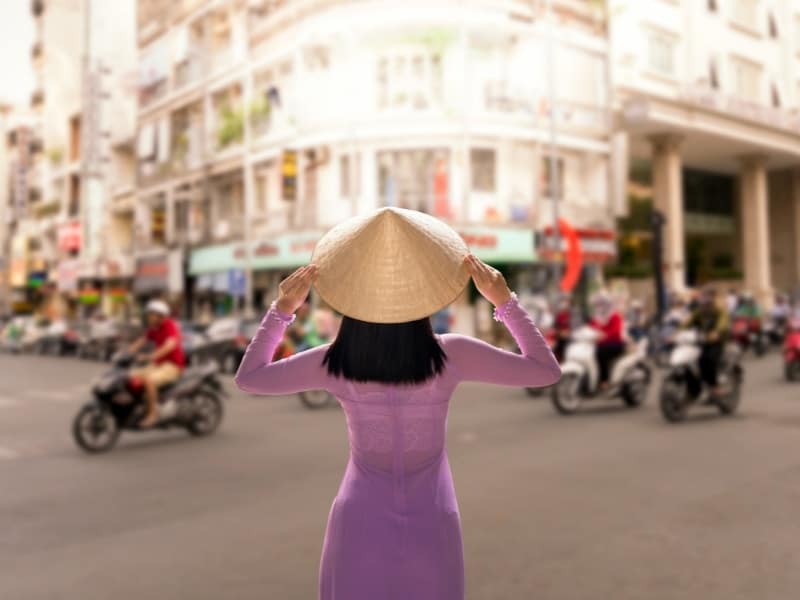 View from behind of woman wearing pink traditional clothing and straw hat while facing a busy intersection in Ho Chi Minh city