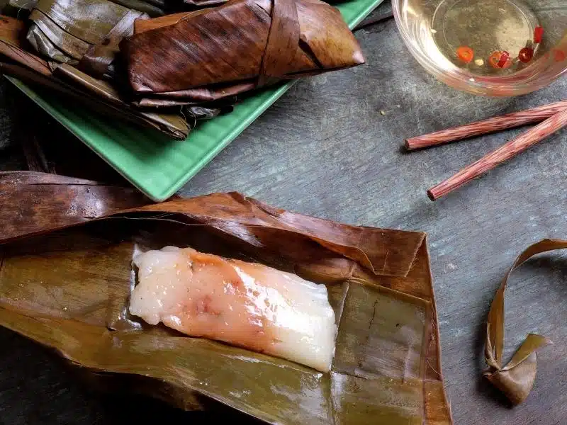 A Vietnamese banh nam with the banana leaf open to see the snack inside with some dipping sauce in a small bowl above it