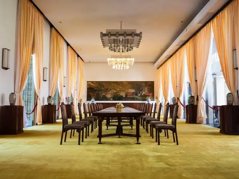 Inner meeting room at the Independence Palace in Saigon, with a long table and wooden chairs, and large curtains on the windows on either side