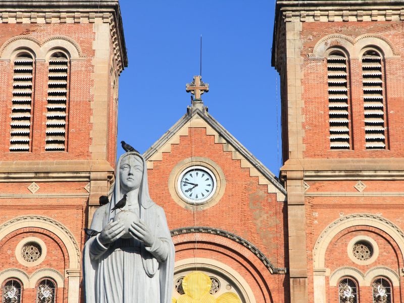 Close up of a gray statue of a woman with a black bird on its head and a pink Vietnamese cathedral behind