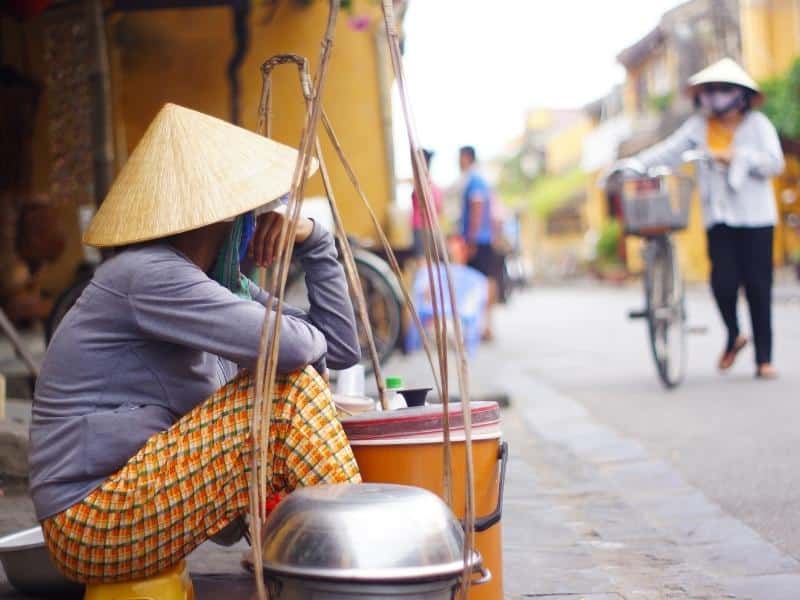 A Vietnamese vendor on the street in Vietnam wearing a rice hat, and another Vietnamese person approaching with a bicycle and also wearing a rice hat