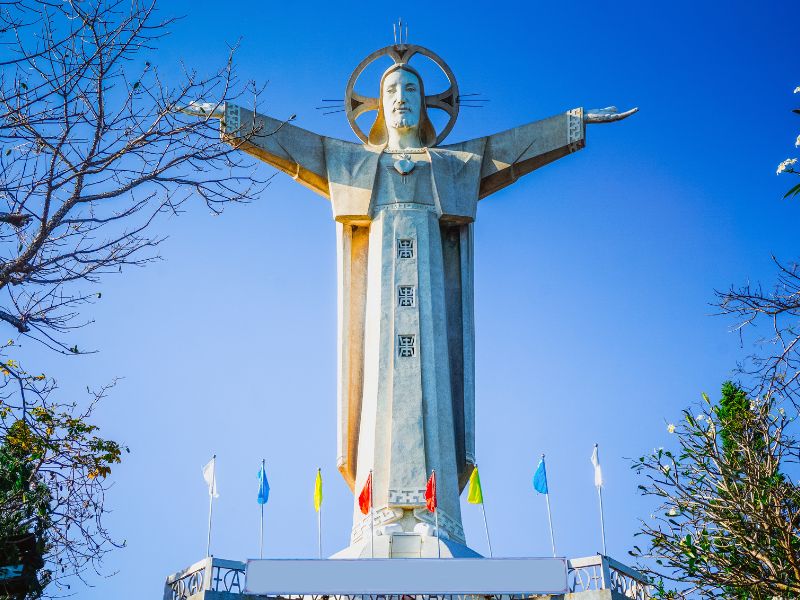 A white Jesus statue in Vietnam with arms outstretched, colorful flags at the bottom, and tree branches on the sides