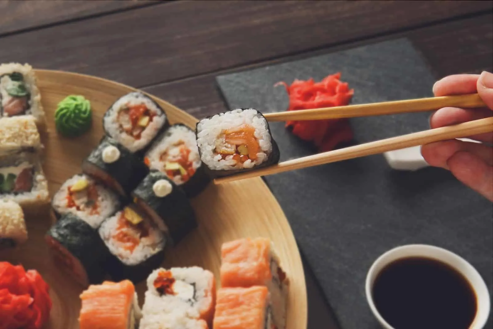 Japanese food restaurant, sushi maki roll plate, platter set. Hand take roll. Set for two with chopsticks, ginger, soy, wasabi. Sushi at rustic wood background and black stone. POV at black. Closeup.